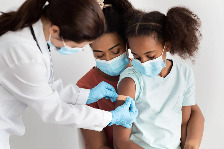 A girl sits on her mother's lap while a nurse puts a plaster on her arm