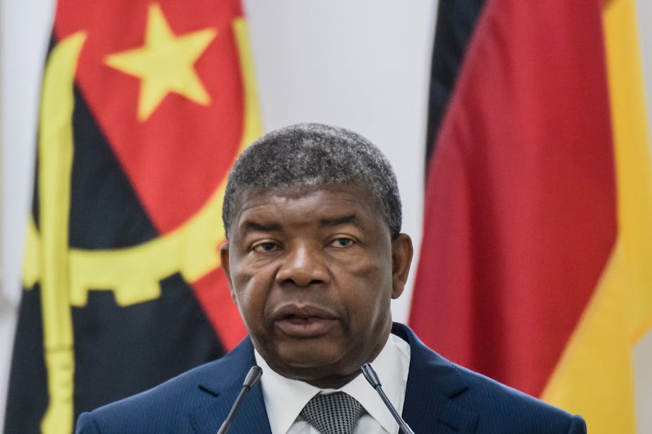 A man wearing a suit and tie speaks into two small microphones at a podium, in front of the flags of Angola and Germany behind him.