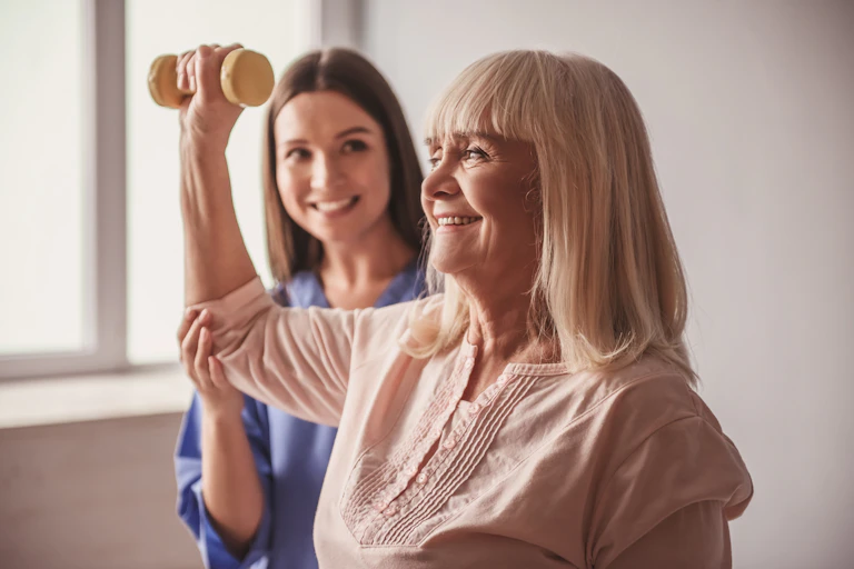 A young female nurse helping an older woman while she lifts a weight.
