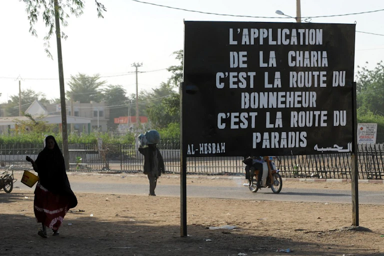 Panneau stipulant que "L'application de la Charia, c'est la route du bonneheur, c'est la route du paradis" au milieu d'un trottoir de terre battue avec une route derrière et une femme en robe noir à sa gauche tenant un seau jaune.