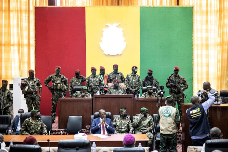 Men in military uniform sit and stand in front of a red, yellow and green flag
