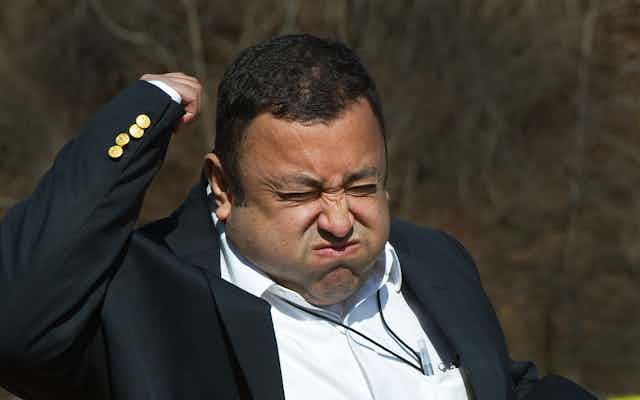 A man wearing a blazer, holding a microphone in his left hand, raising his right arm and squeezing his eyes shut