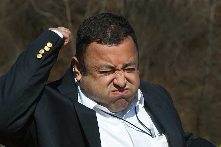 A man wearing a blazer, holding a microphone in his left hand, raising his right arm and squeezing his eyes shut