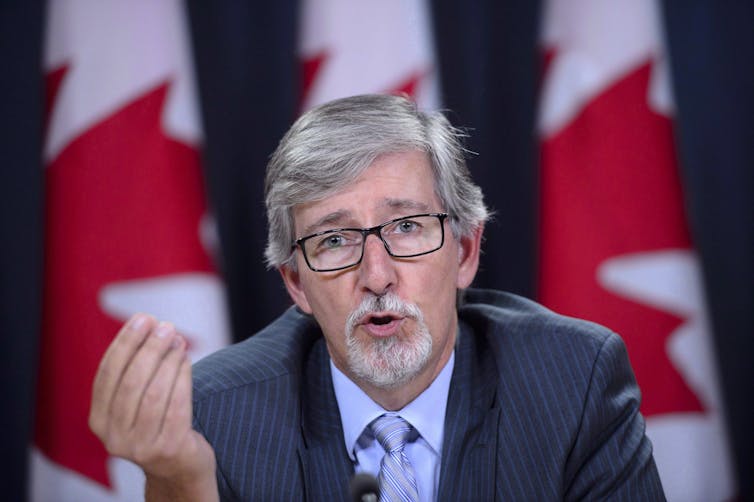 privacy commissioner Daniel Therrien sits at a news conference making a hand gesture with Canadian flags in the background