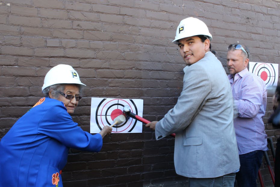 An elder and a young man are seen wearing hard hats smiling and swinging a hammer against a brick wall.