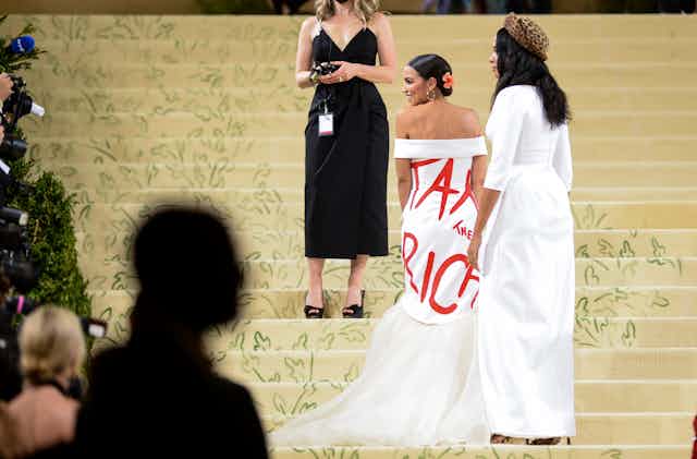 Congresswoman Alexandria Ocasio-Cortez  walks up the stairs at the Met Gala wearing a white dress with "Tax the Rich" written on it.