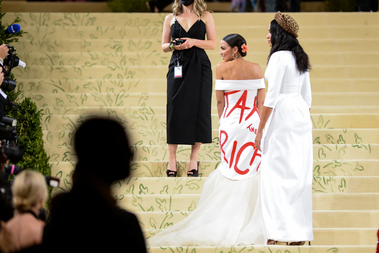 Congresswoman Alexandria Ocasio-Cortez walks up the stairs at the Met Gala wearing a white dress with "Tax the Rich" written on it.