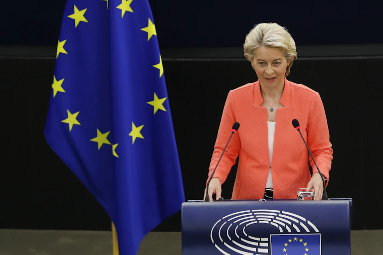 European commission president Ursula von der Leyen stands behind a podium, wearing an orange jacket. There is an EU flag beside her.