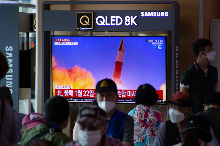 People with masks watch a TV screeen showing the launch of a ballistic missile