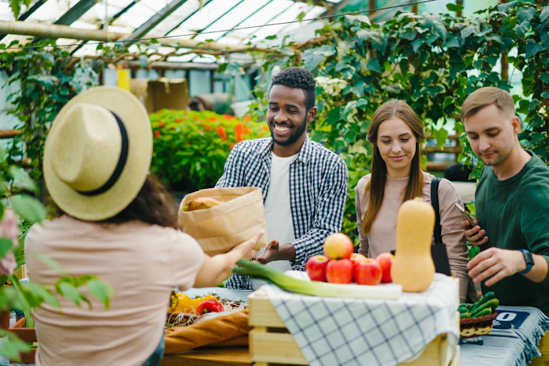 A group of friends purchasing vegetables from a woman at a farmer's market, inside a greenhouse