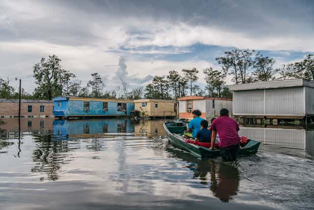 A family in a small motorboat goes through a flooded street among trailer homes.