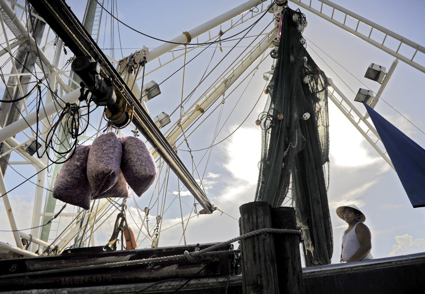 A fisherman in a round hat watches stands on a boat with rigging as bags of shrimp being moved off by a boom arm.
