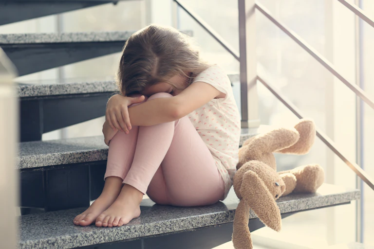Une fillette triste se cache la tête, assise dans les escaliers, un toutou à ses côtés