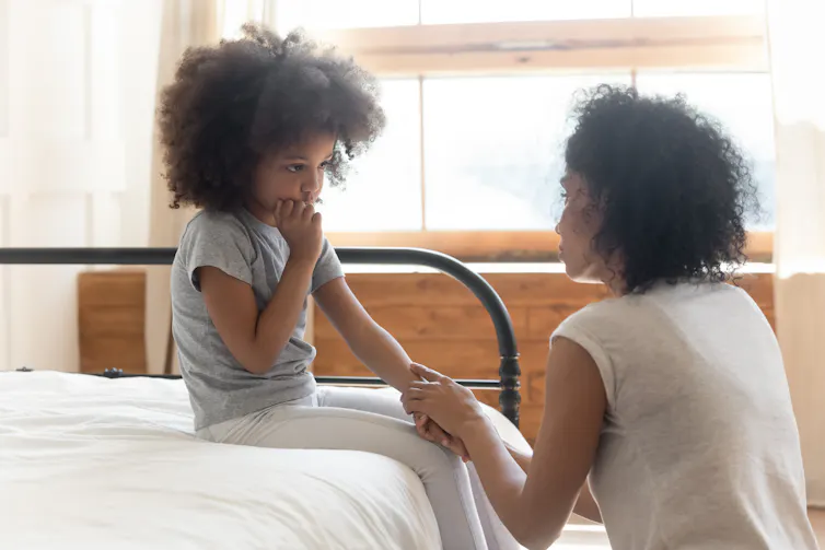 Une fillette assise sur un lit, avec sa mère qui lui prend la main