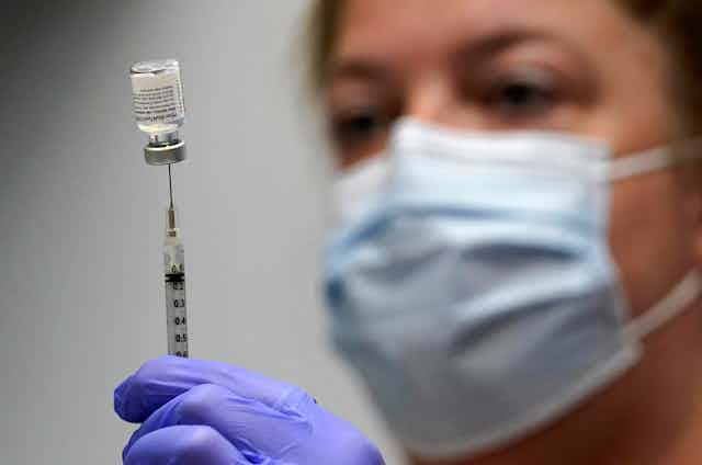 A pharmacy technician wearing a mask looks on as she loads a syringe with COVID-19 vaccine.