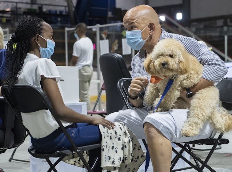A man in a mask holds a dog in front of a young woman, also wearing the mask