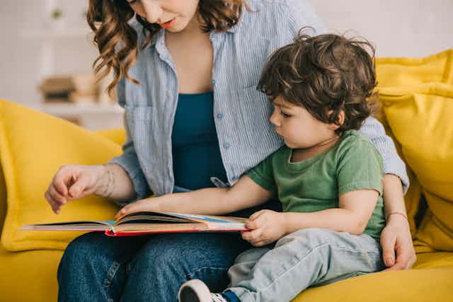 A toddler reads a book with their mother while sitting on an orange couch.