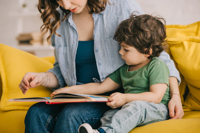 A toddler reads a book with their mother while sitting on an orange couch.