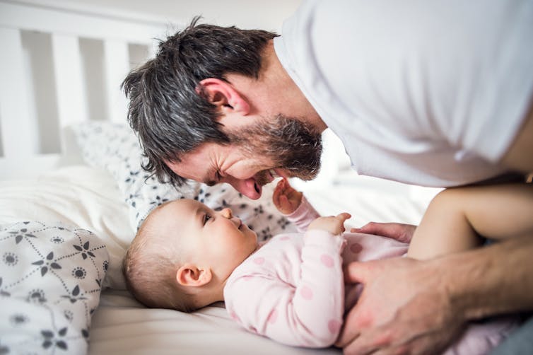 A man talks to his baby who lays on a bed.