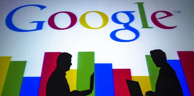 The silhouettes of two people using laptops are seen against the Google logo.