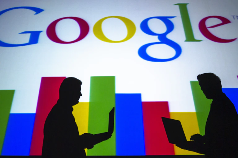 The silhouettes of two people using laptops are seen against the Google logo.
