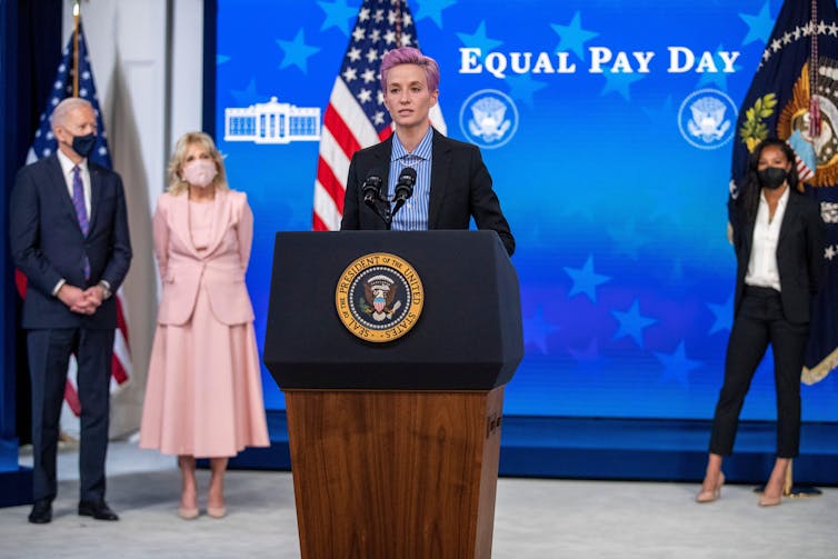 Megan Rapinoe, with short, purple hair and wearing a suit, speaks at a podium with the White House insignia. A background image reads 'Equal Pay Day', and President and First Lady Joe and Jill Biden stand behind Rapinoe.