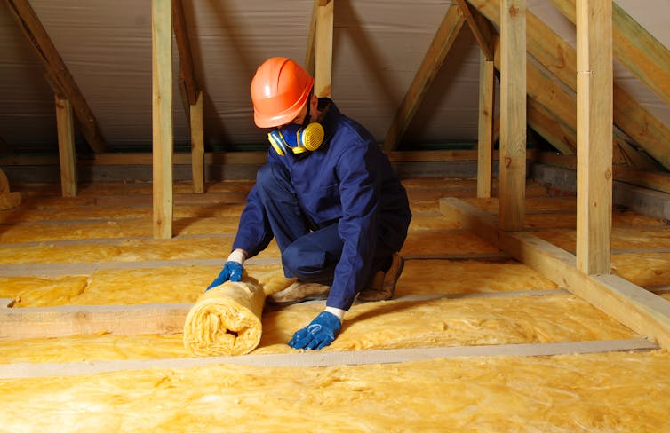 A worker in blue overalls rolls out wool in an attic.