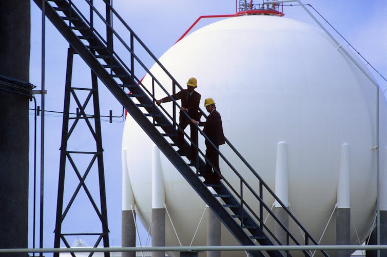 Two workers climb stairs with a large storage tank in the background.