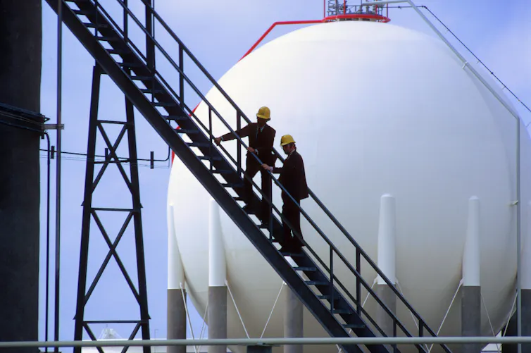 Two workers climb stairs with a large storage tank in the background.