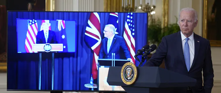Joe Biden stands at a podium with Boris Johnson and Scott Morrison on screens beside him.