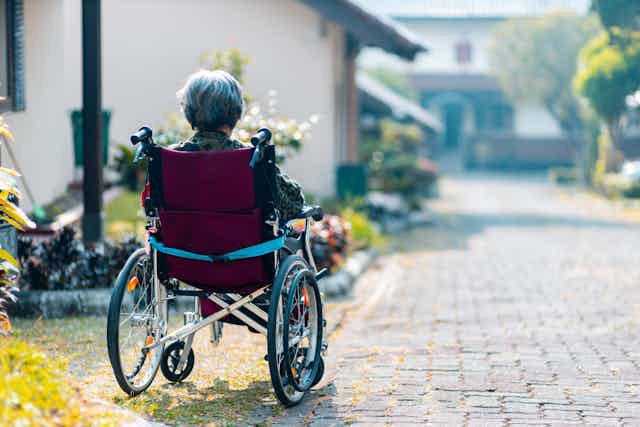 Photo d’une dame âgée dans un fauteuil roulant, de dos, au bord d’une allée dans un jardin.