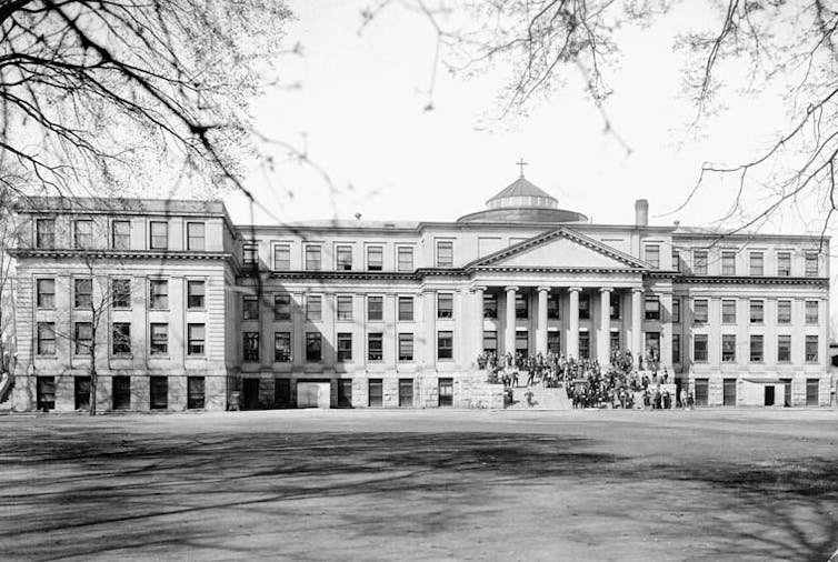 People standing on steps in front of a university building set back from a large grassy area in a black and white photo.