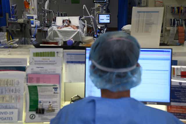 An ICU nurse sits at a computer while a patients lays in bed in the background.