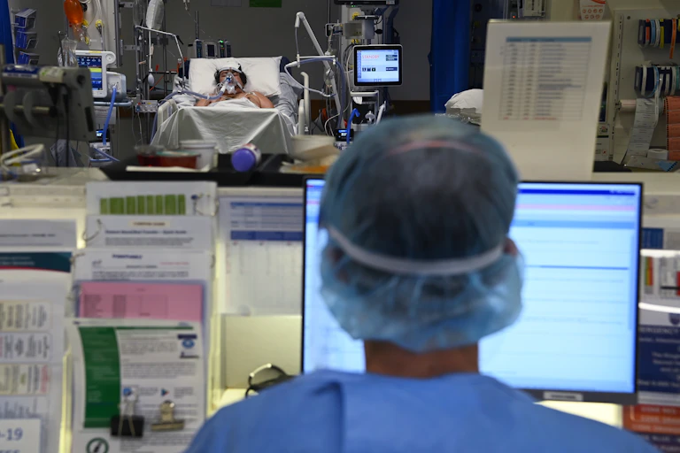 An ICU nurse sits at a computer while a patients lays in bed in the background.