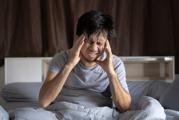 Man sits up in bed, scrunching has face and cradling his head, with a headache.