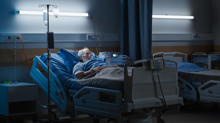 An elderly man with a breathing mask lays in a hospital bed in a dark ward room.