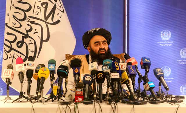 Afghanistan's foreign minister, Amir Khan Muttaqi, sits at a desk surrounded by press microphones and with a Taliban flag in the background.