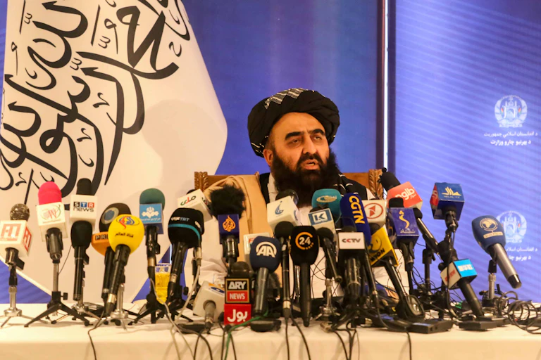 Afghanistan's foreign minister, Amir Khan Muttaqi, sits at a desk surrounded by press microphones and with a Taliban flag in the background.