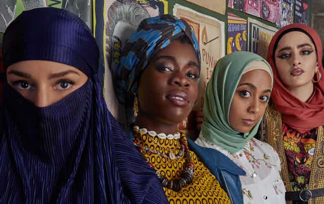 Five girls of diverse ethnicity, most wearing head coverings, stand by a wall.