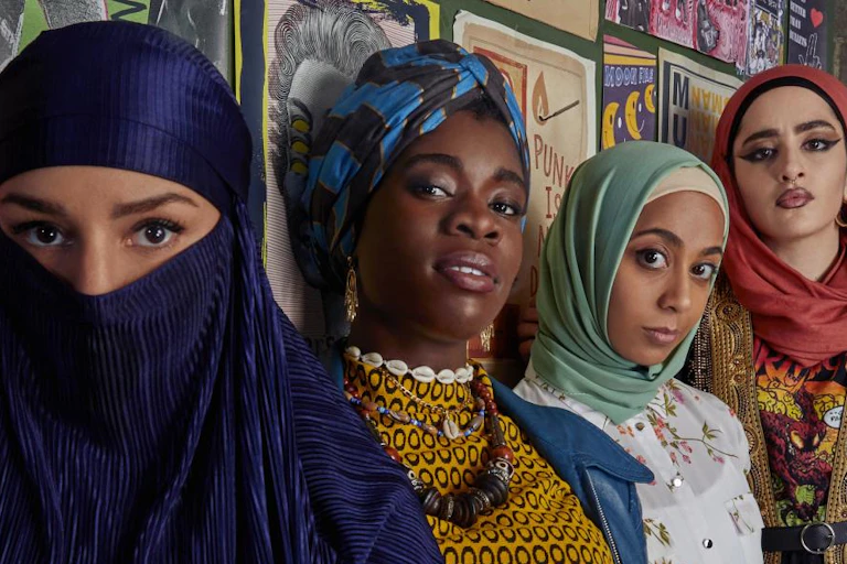 Five girls of diverse ethnicity, most wearing head coverings, stand by a wall.