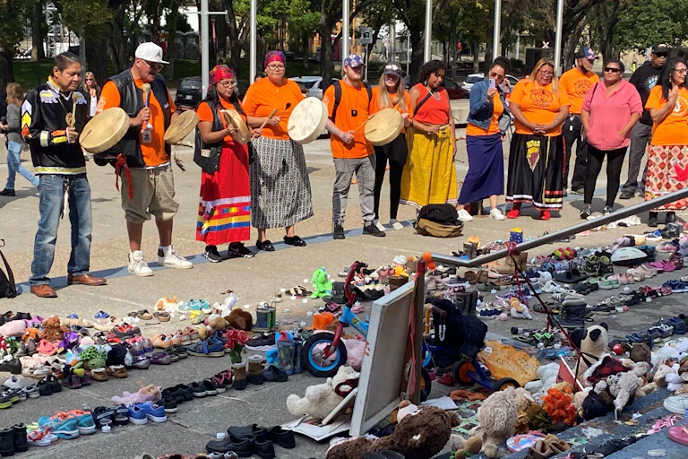 A group of people wearing orange hold hand drums in front of hundreds of shoes on steps
