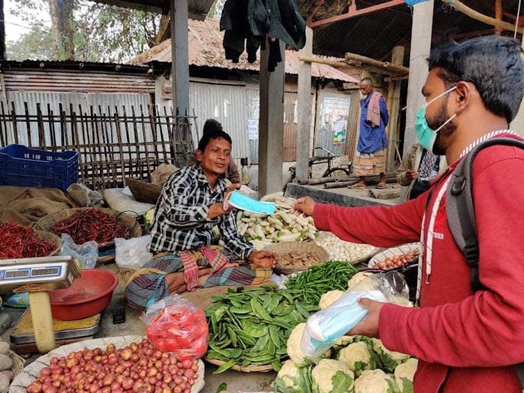 A man wearing a surgical mask handing a mask to a woman working at a vegetable stand.