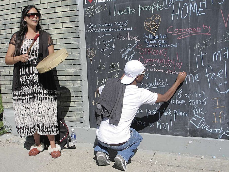 A woman drums while a young man writes on a chalk wall.