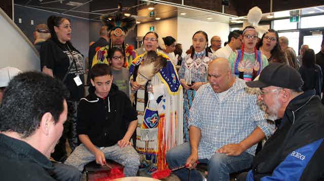 People are seen standing around a drum in regalia or seated and drumming