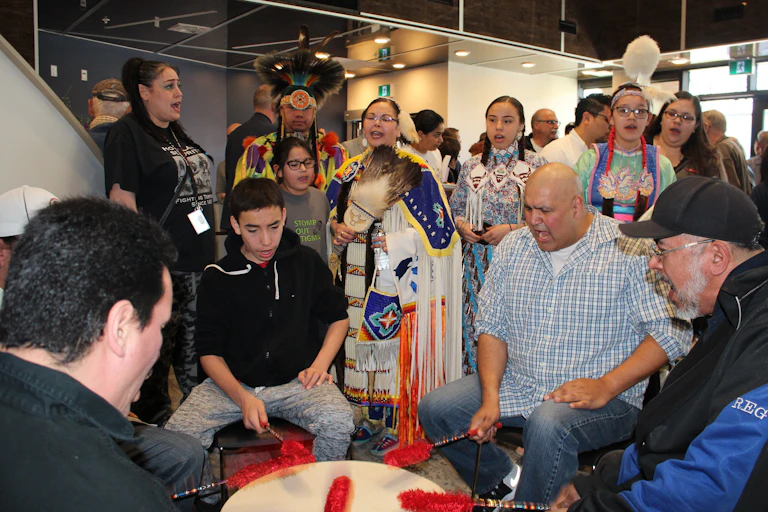 People are seen standing around a drum in regalia or seated and drumming