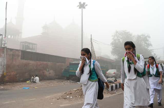 Three girls walk through smoggy conditions, two holding handkerchiefs over the noses.
