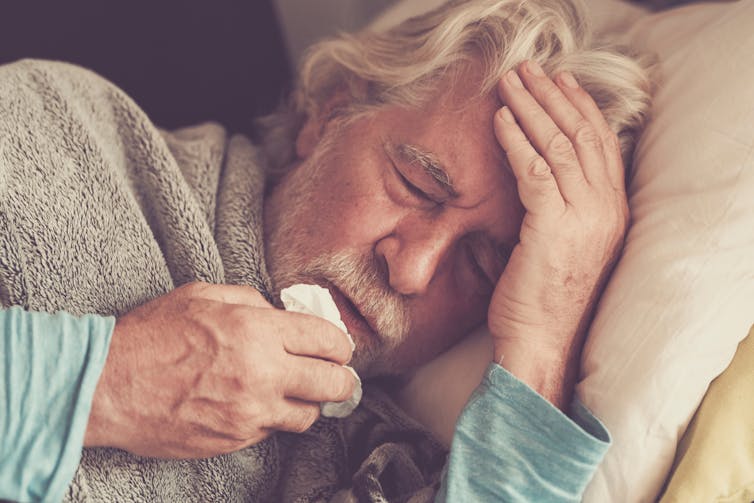 Older man with cold symptoms lays down, wrapped in a blanket, cradling his head, holding a tissue to his nose.