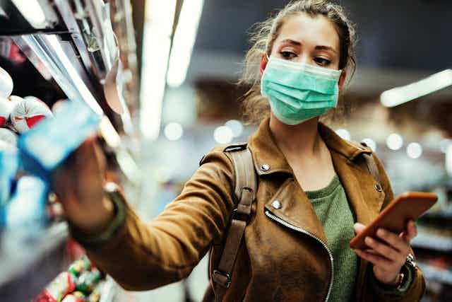 Woman in a mask and leather jacket shops for groceries while holding her smartphone.