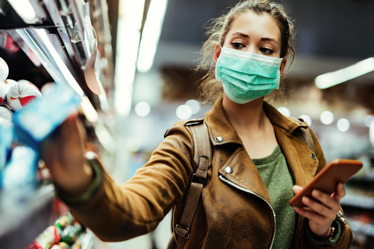 Woman in a mask and leather jacket shops for groceries while holding her smartphone.