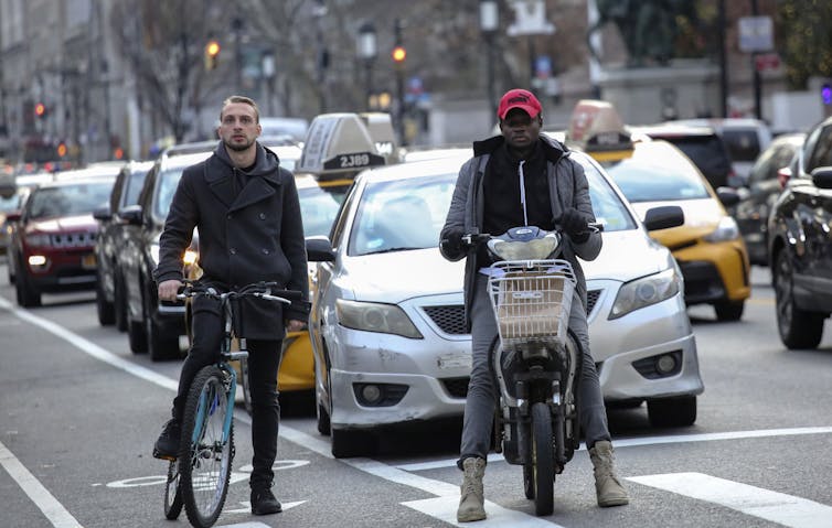 Two men on bicycles wait with taxis at a city stoplight.